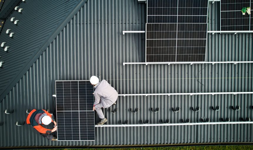 Mounters building photovoltaic solar module station on roof of house. Men electricians in helmets installing solar panel system outdoors. Concept of alternative and renewable energy. Aerial view.